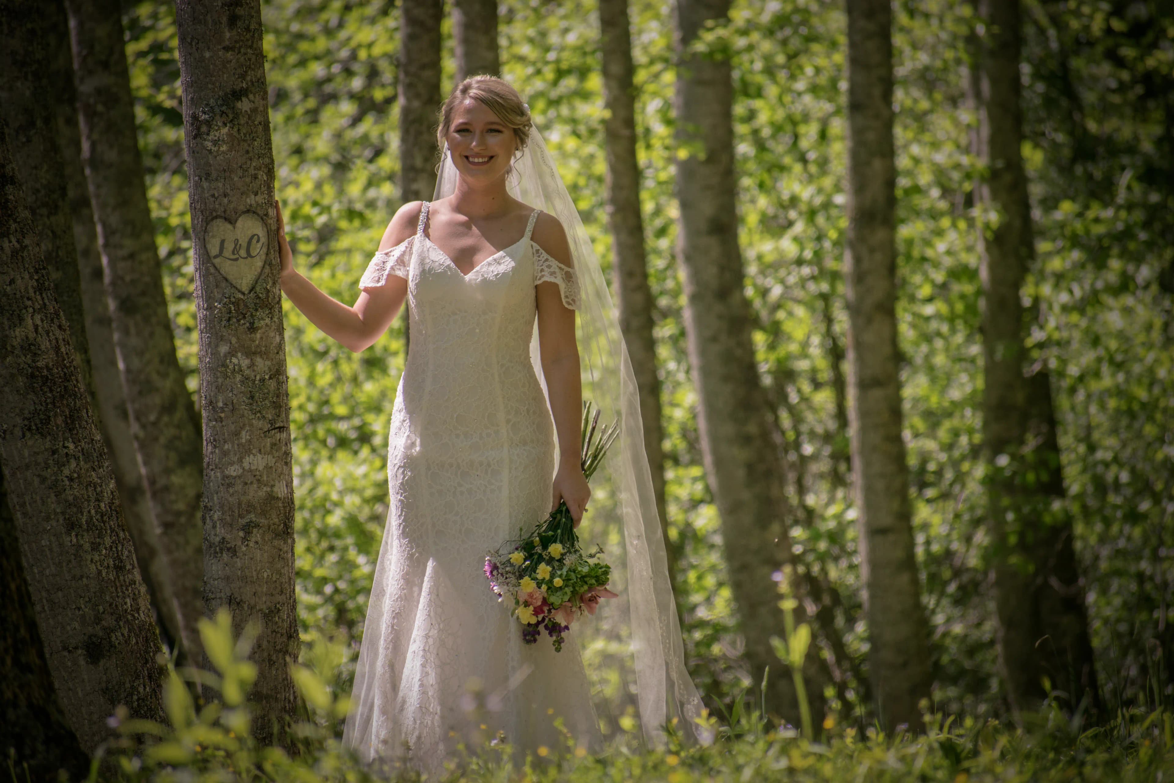 The bride smiling at the camera with her hand on a tree showing the initials of the couple.