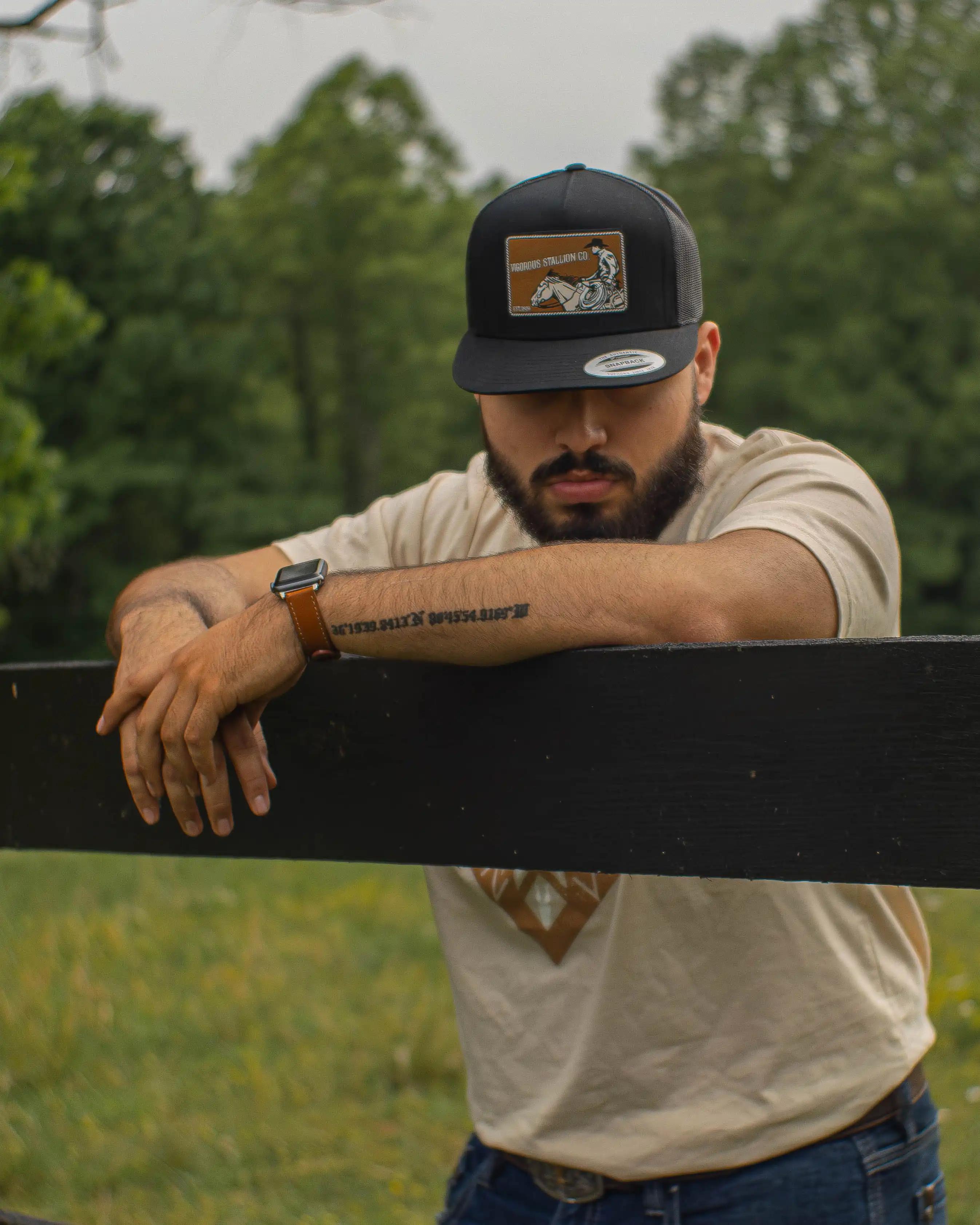 A person leaning onto a black wood fence wearing the Traveler snapback hat and the Aztec Sand tan-colored shirt.
