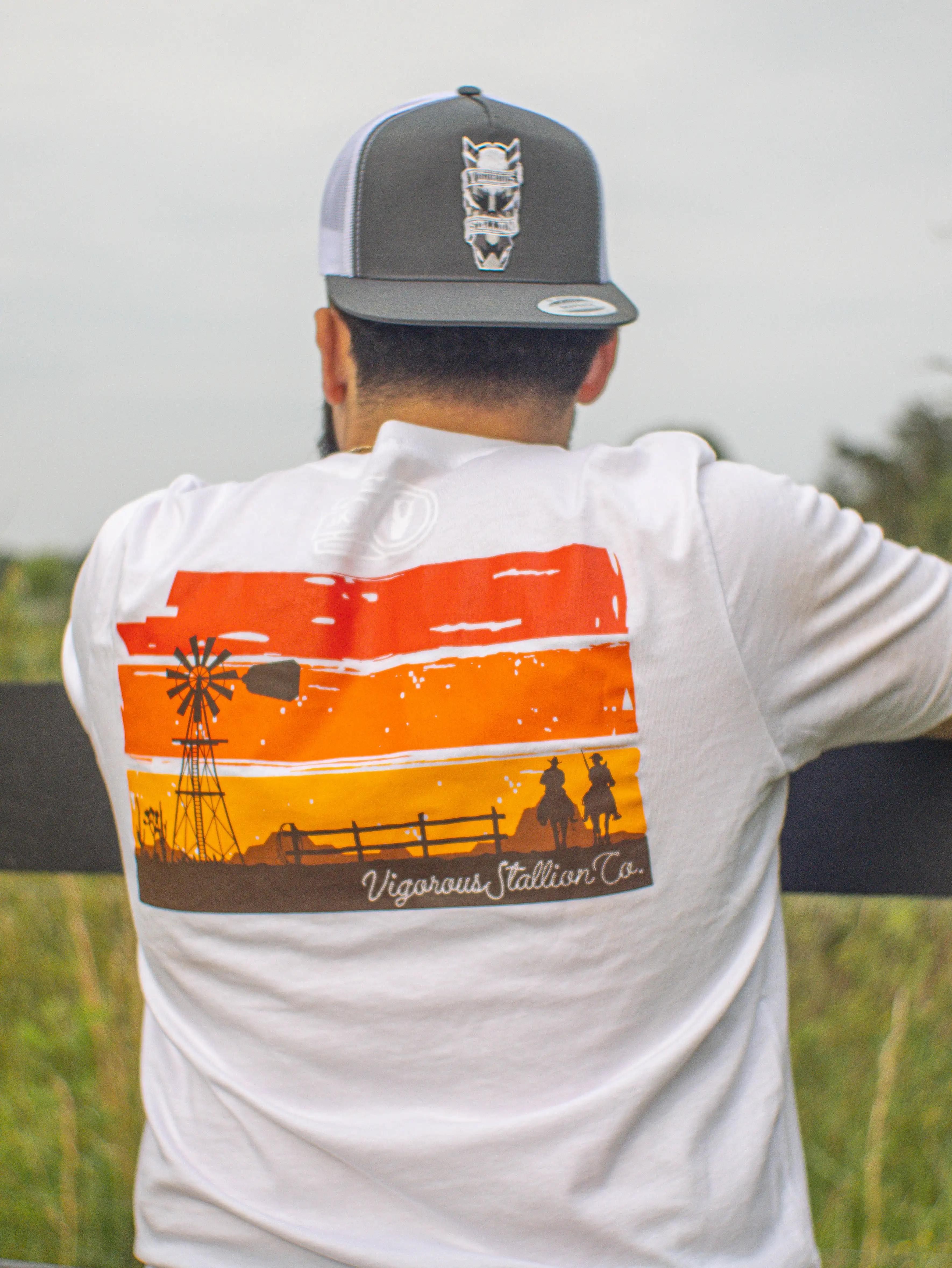 A person leaning onto a black wood fence photographed from the back wearing the Aztec Stallion snapback hat and the Sunset Bandit white shirt.
