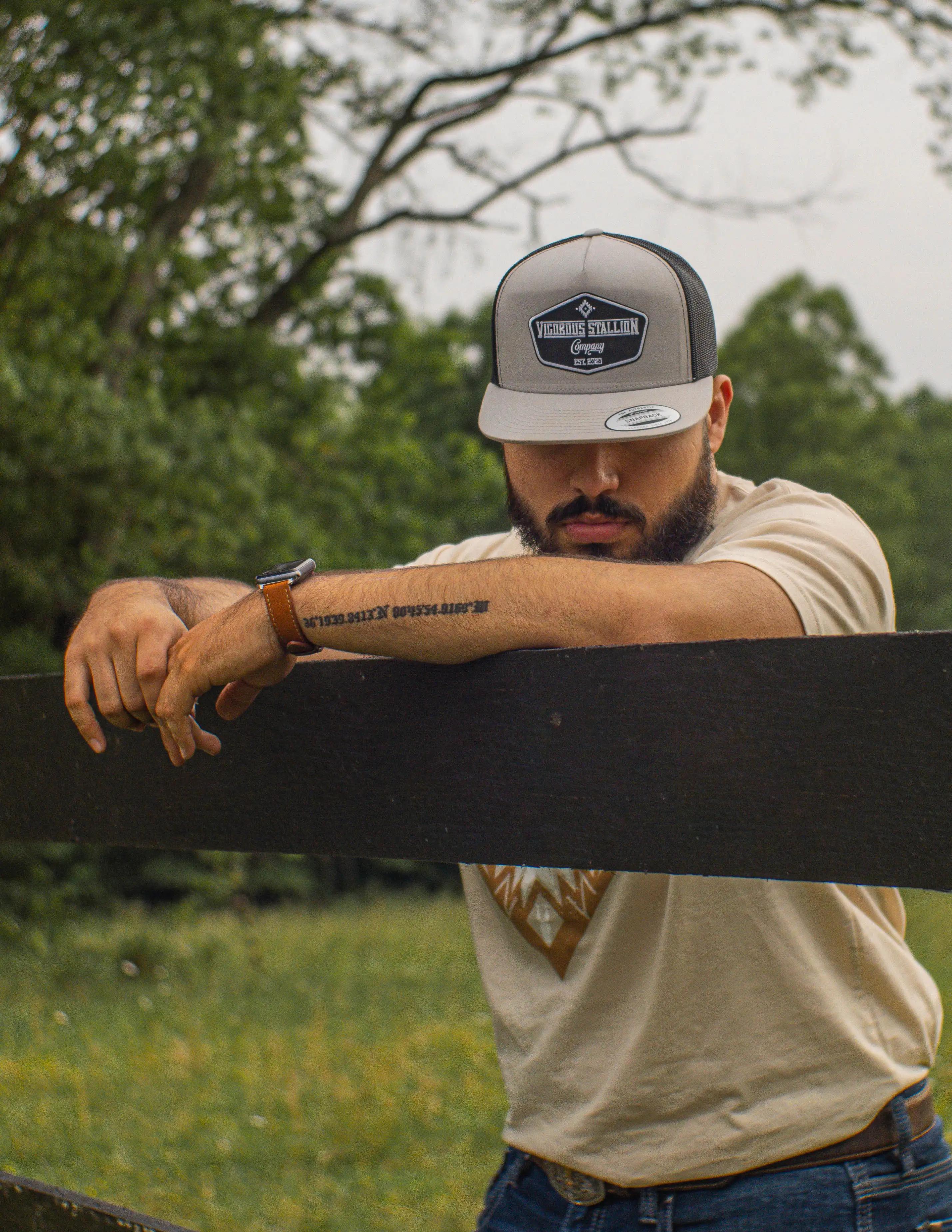 A person leaning onto a black wood fence wearing the Classic snapback hat and the Aztec Sand tan-colored shirt.