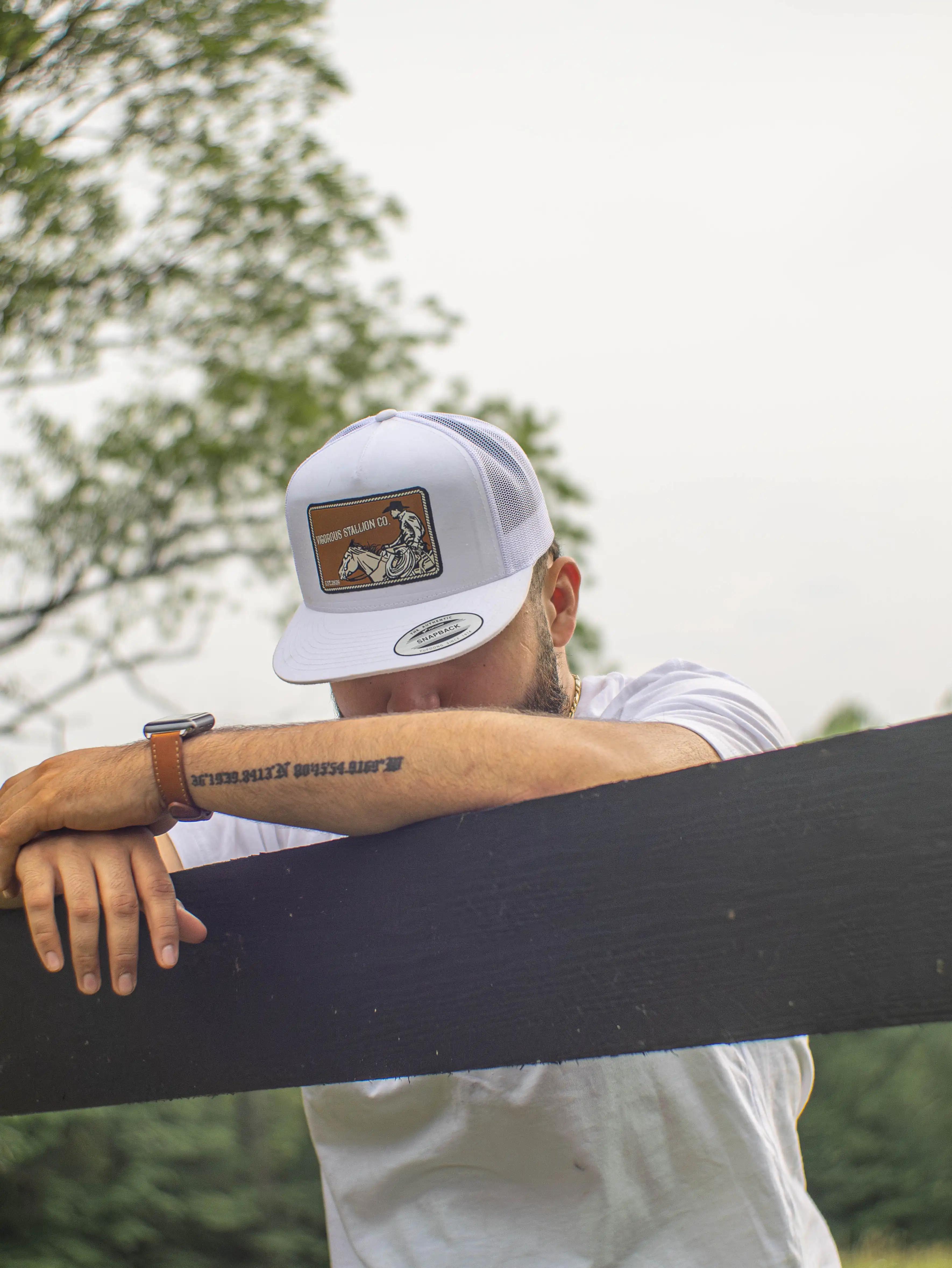 A person leaning onto a black wood fence wearing the Roper snapback hat and the Sunset Bandit white shirt.