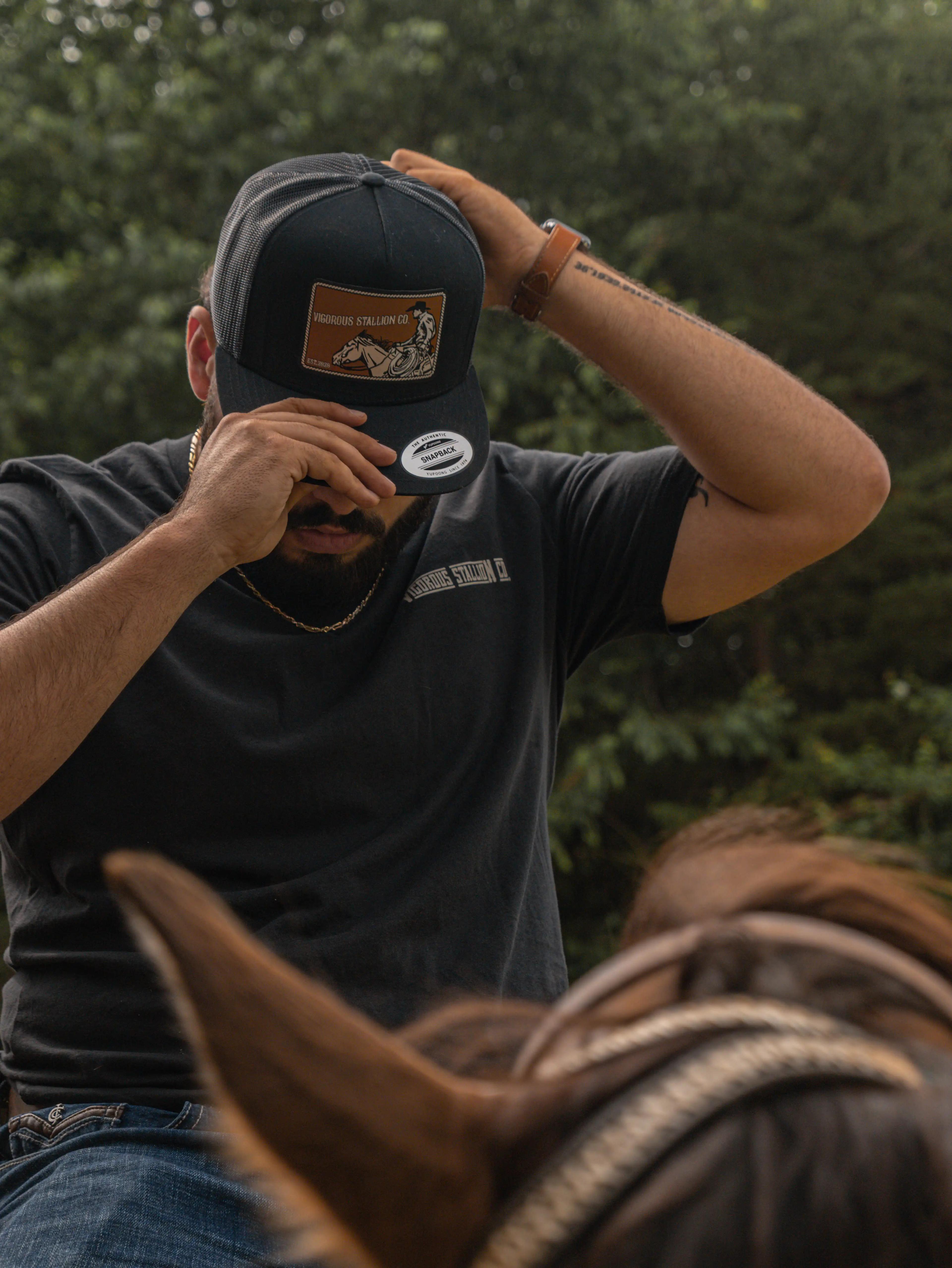A person on top of a horse adjusting the snapback Traveler hat wearing the Black Stallion shirt.