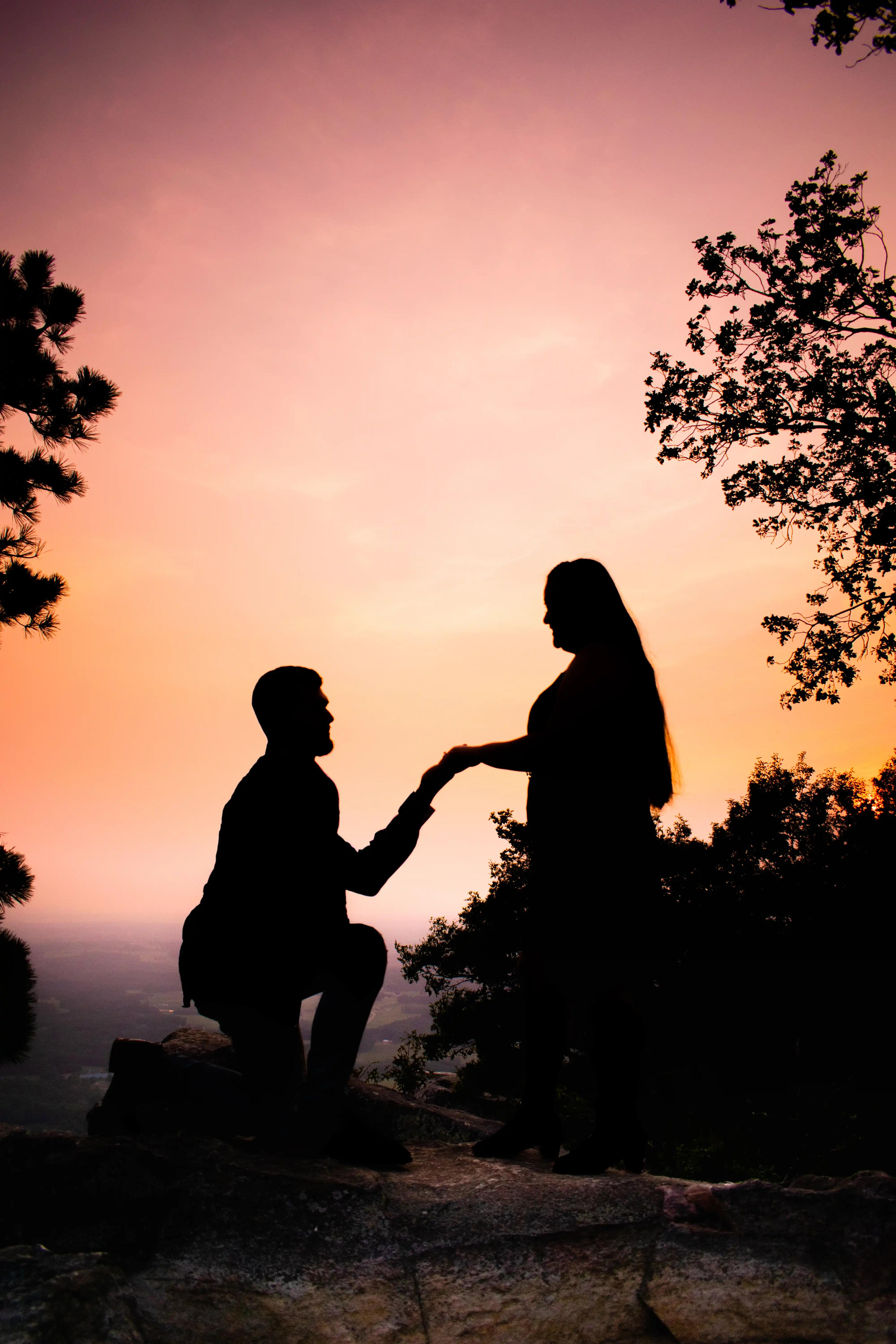 the groom silhouetted by the sunset getting down on one knee to propose to his future wife.