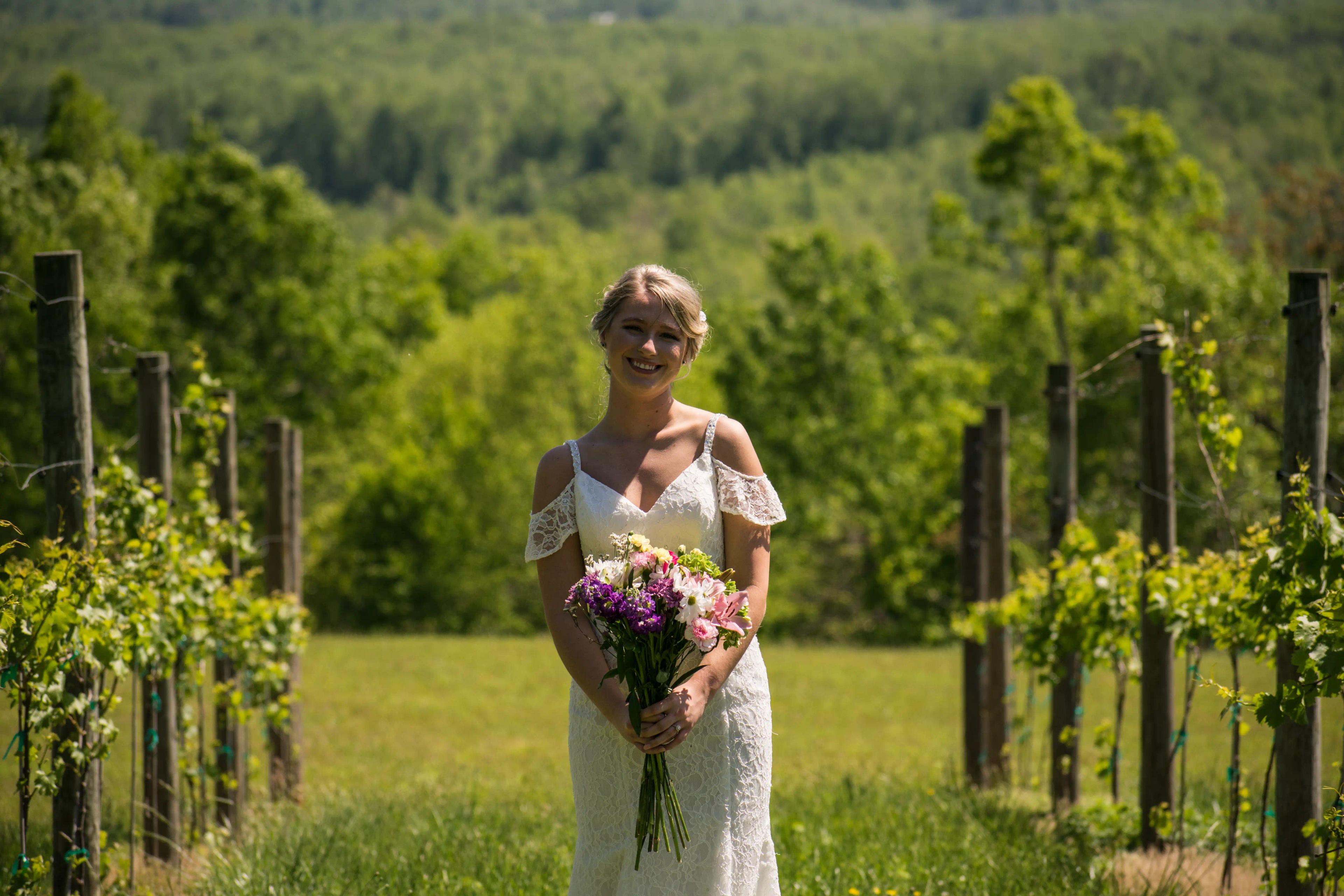 The bride standing in the middle of a vineyard with grape vines on either side with the bride in the middle smiling.