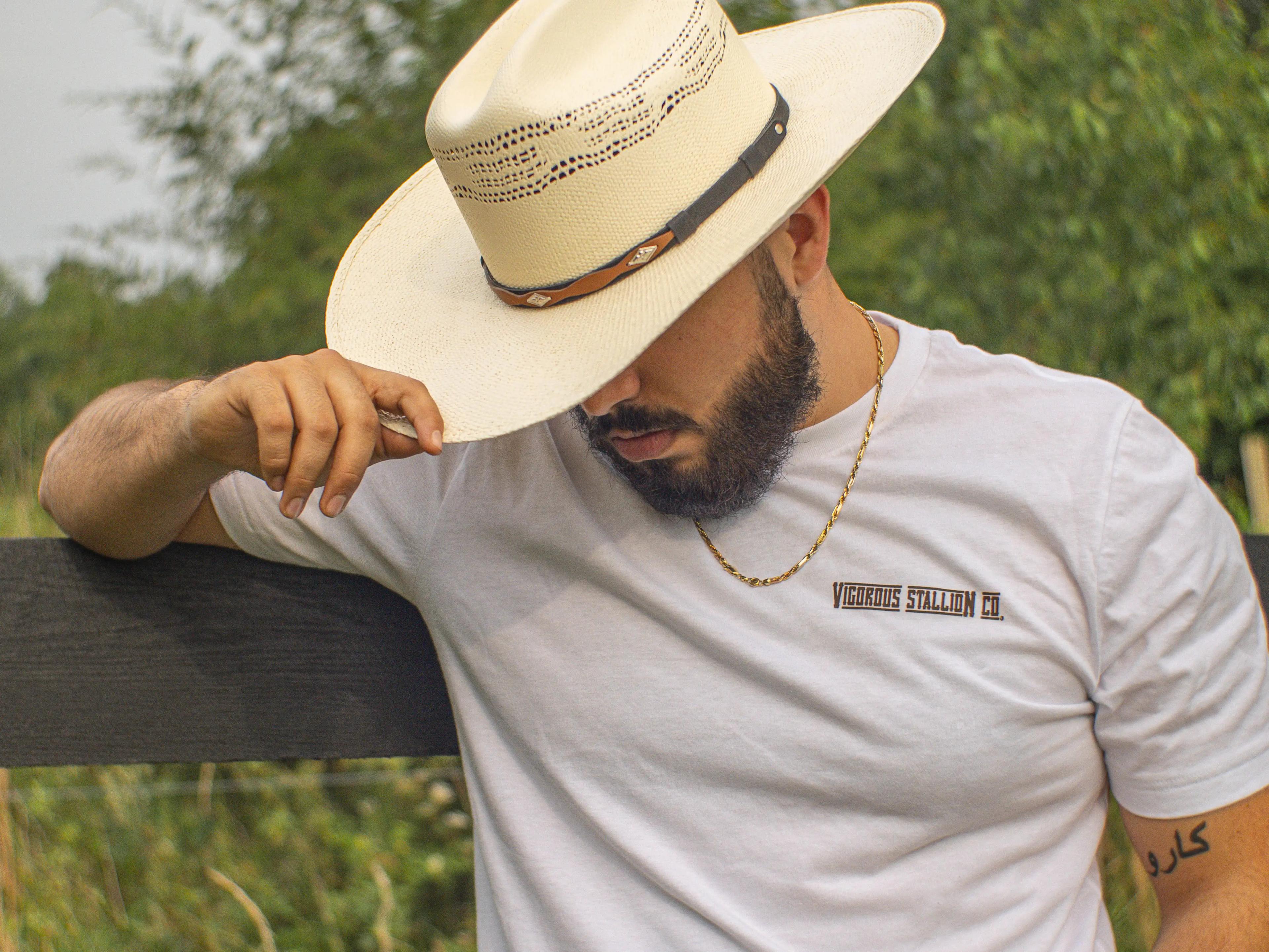 A person leaning onto a black wood fence wearing a tan cowboy hat and the sunset bandit white shirt.